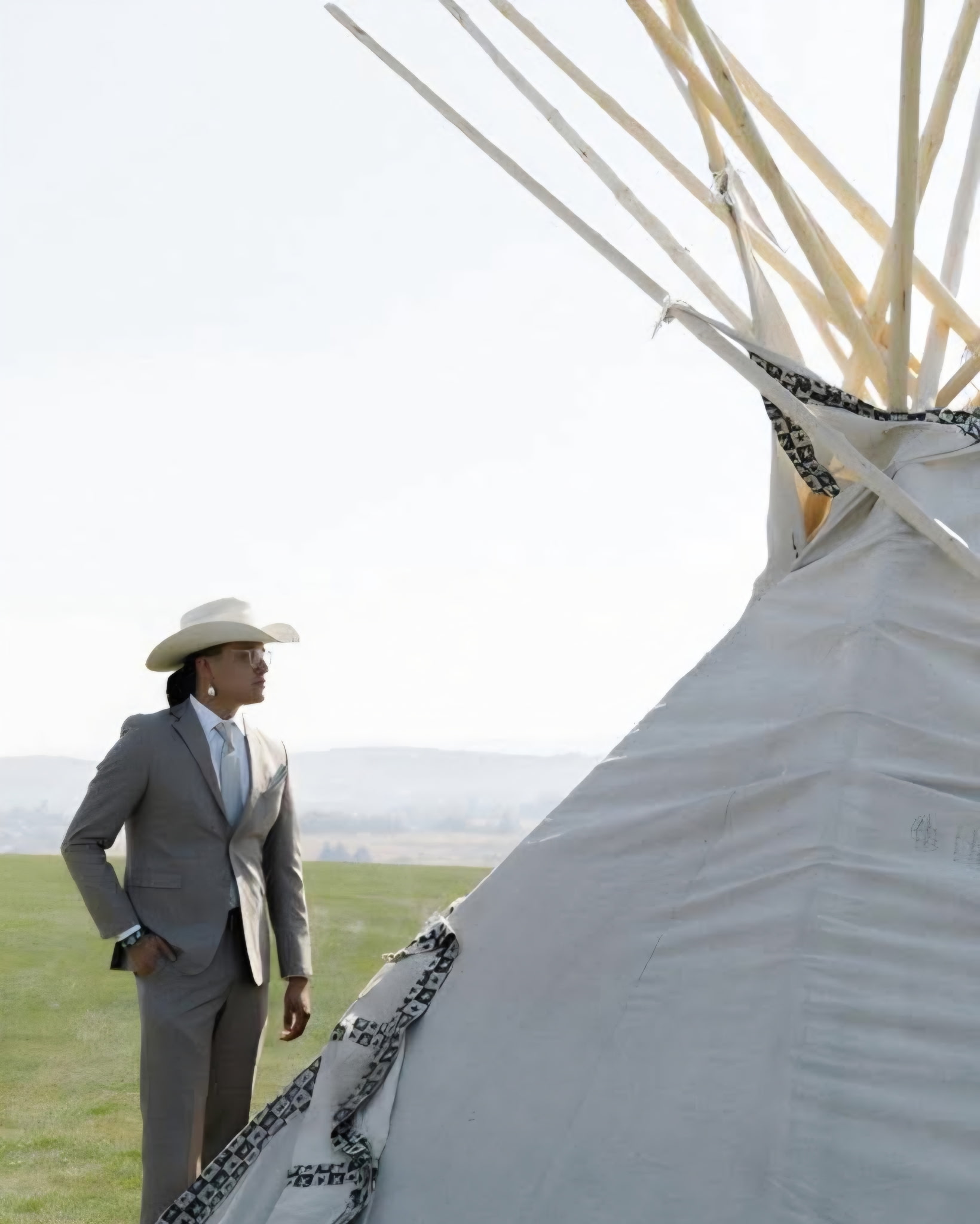 A person in a light-colored suit and cowboy hat standing beside a tipi in an open field